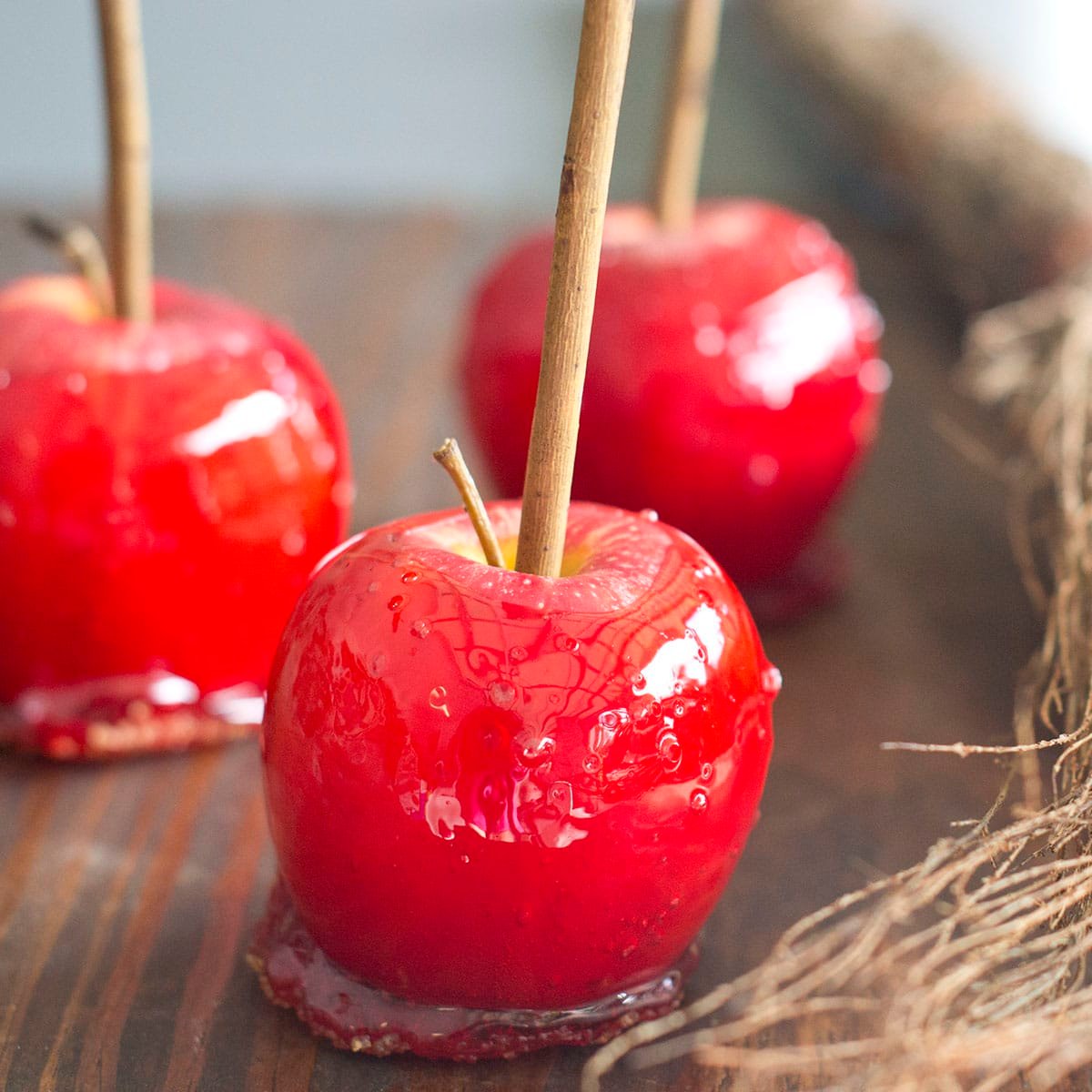 Three red apples coated in candy with rustic sticks coming out of the tops. There's grassy hay beside them.