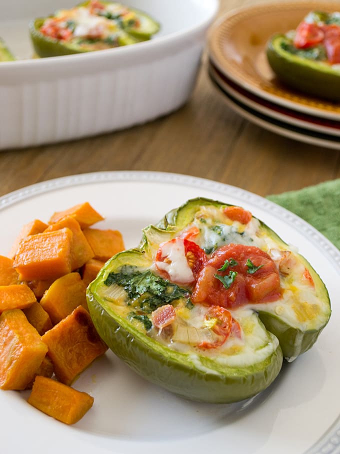 White round plate with half a baked green bell pepper stuffed with kale, onion, bacon, and tomato topped with melted mozzarella cheese. Next to the pepper on the plate is cubed, baked orange sweet potato chunks. In the background is a rectangular casserole dish with another half a stuffed pepper in it. Next to the casserole dish is a stack of brown plates also with a half of a stuffed pepper on the top plate.