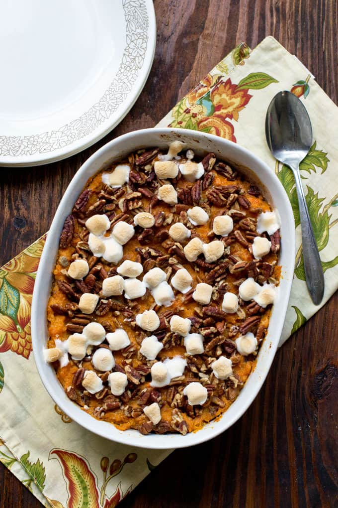 white casserole dish with mashed sweet potatoes, toasted marshmallows and chopped pecans; cream colored cloth with yellow, red, and orange flowers under dish with spoon setting on cloth; white plates with a light brown flower design around edge in corner