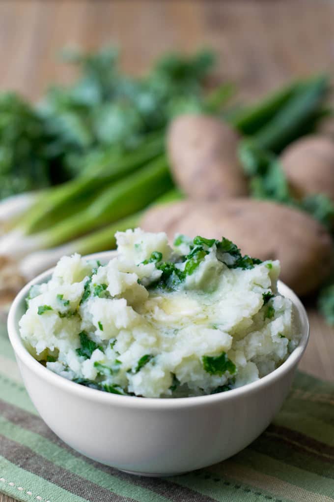 White bowl sitting on a black and green striped dish towel. The bowl is filled with mashed potatoes with cooked kale mixed in. A melted pat of butter is on top of potatoes.
