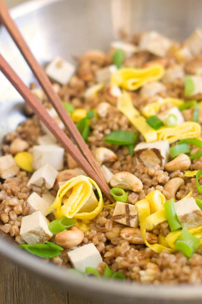 close up of fried farro, done in the style of fried rice, with chunks of tofu, eggs, cashews and chopped green onions. Wooden chopsticks are sticking out of the bowl of fried farro.