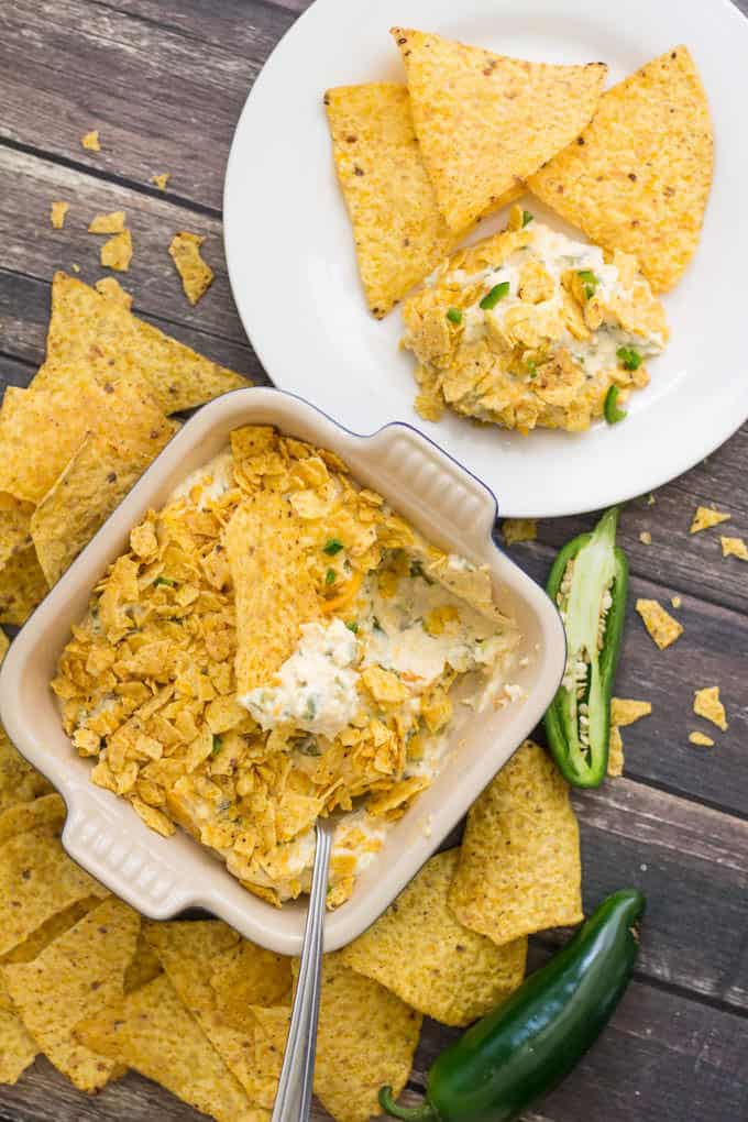 square casserole dish filled with Jalapeño Popper Dip topped with crushed tortillas chips; casserole dish is sitting on a wooden surface with crushed tortillas chips and a half a jalapeño scattered around it; a round white plate with a scoop of dip and some corn chips is above the casserole dish.