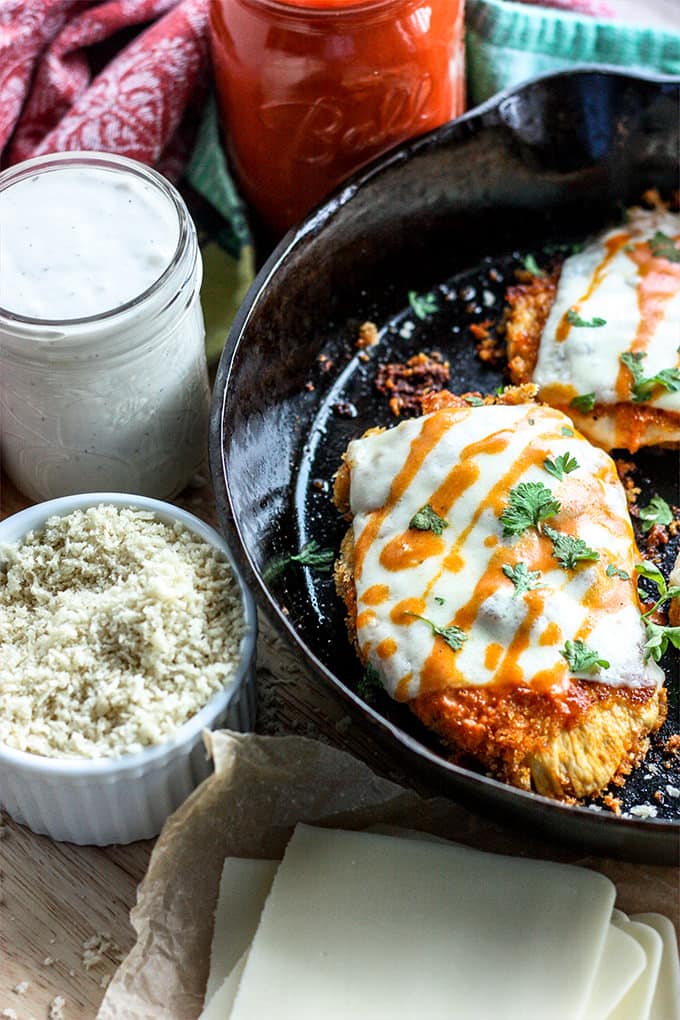 Half of a cast iron skillet with two breaded chicken breasts covered in melted white cheese and drizzles of buffalo sauce on a wood table next to a stack of white cheese slices, a ramekin filled with breadcrumbs and a mason jar filled with a creamy white dressing.