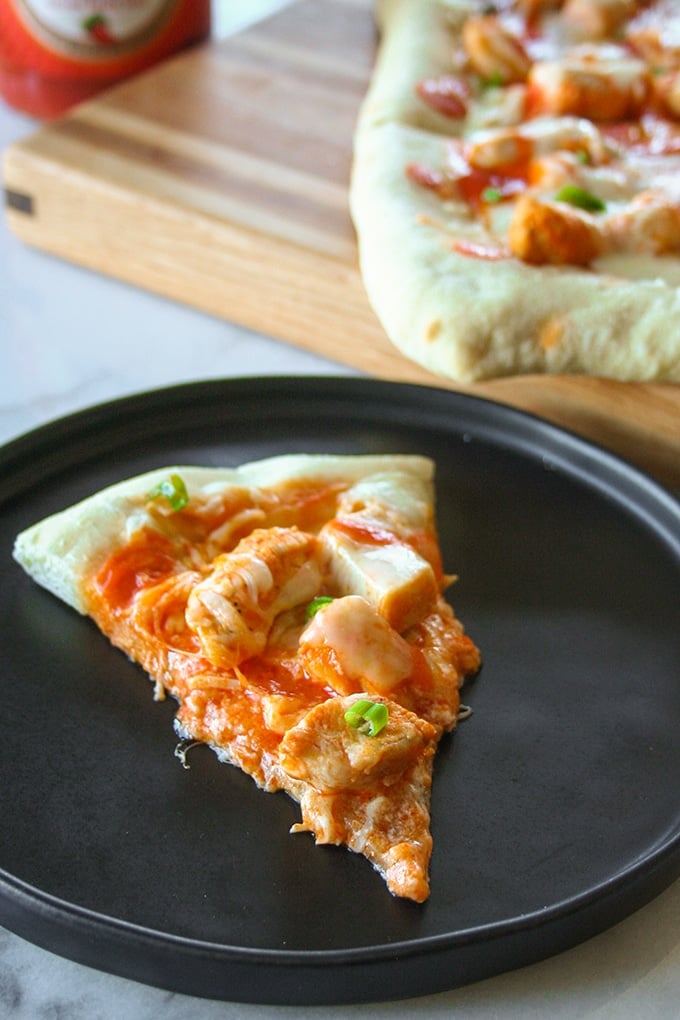 One slice of Buffalo Chicken Pizza sprinkled with green onions on a black round pizza pan. In the background is the corner of a whole pizza on a wooden cutting board.