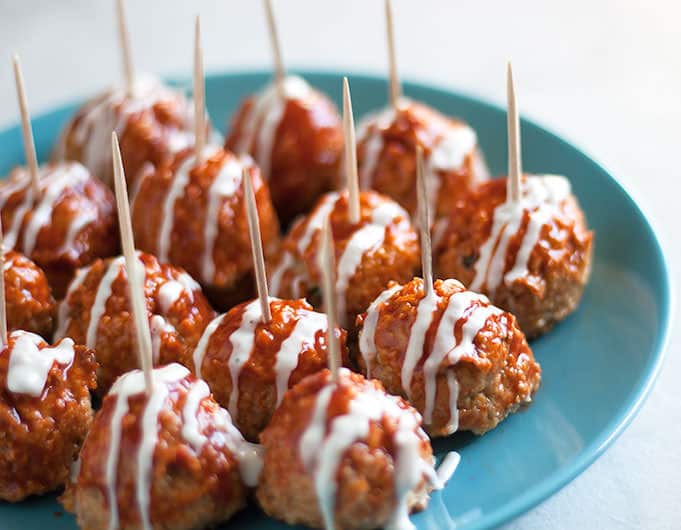 Meatballs on a blue plate with toothpicks sticking out of them, smothered in Buffalo sauce and drizzled with ranch dressing with a glass full of amber beer in the background.