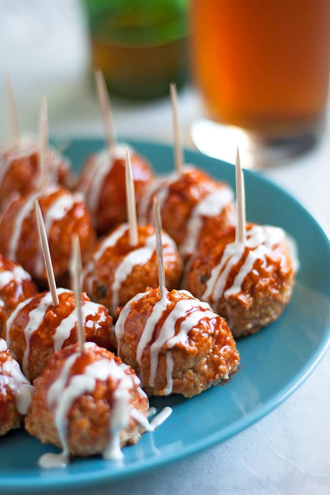 Meatballs on a blue plate with toothpicks sticking out of them, smothered in Buffalo sauce and drizzled with ranch dressing with a glass full of amber beer in the background.