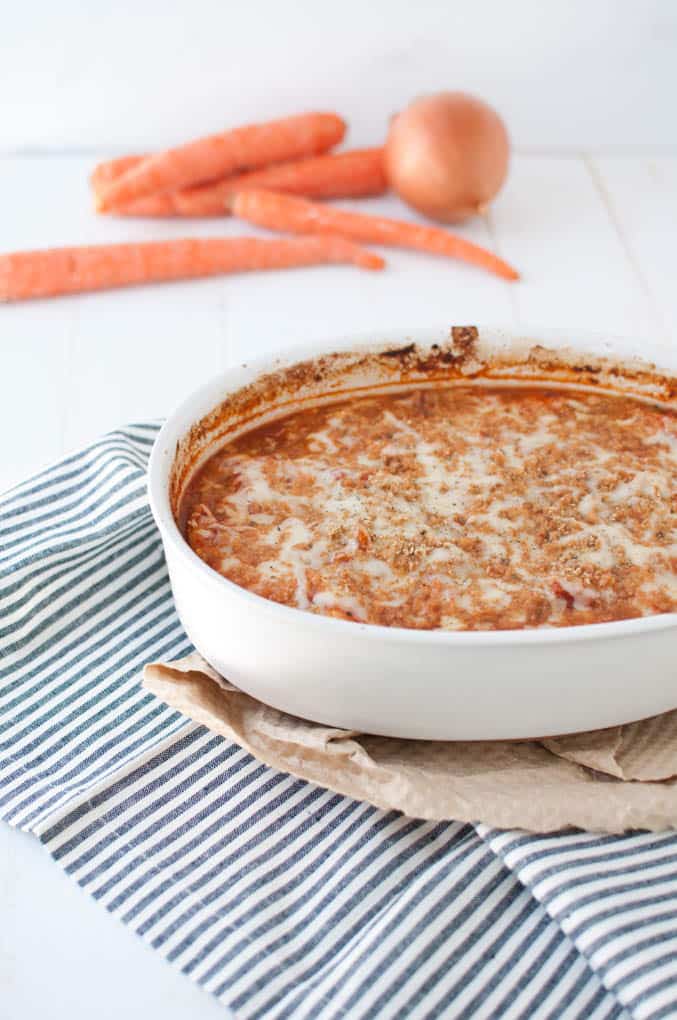 Round casserole dish filled with One Pot Chicken Parmesan that is covered with melted cheese. The dish is sitting on a brown paper towel on a blue and white stripped dish towel on a white  table. In the background is several whole, unpeeled carrots and a whole onion.