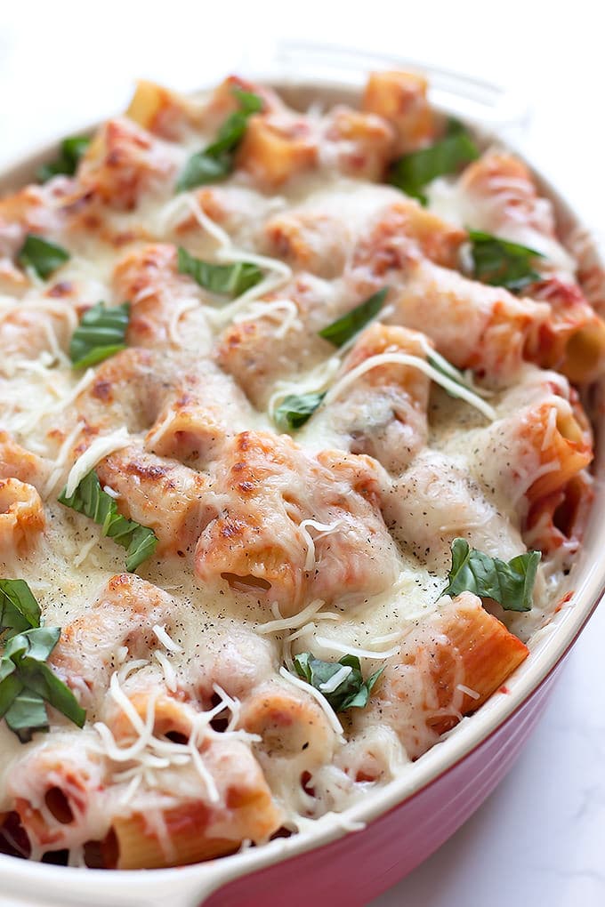 Close up of a red ceramic baking dish filled with a casserole of rigatoni noodles in red sauce with melted white cheese over the top and scattered with roughly chopped basil.