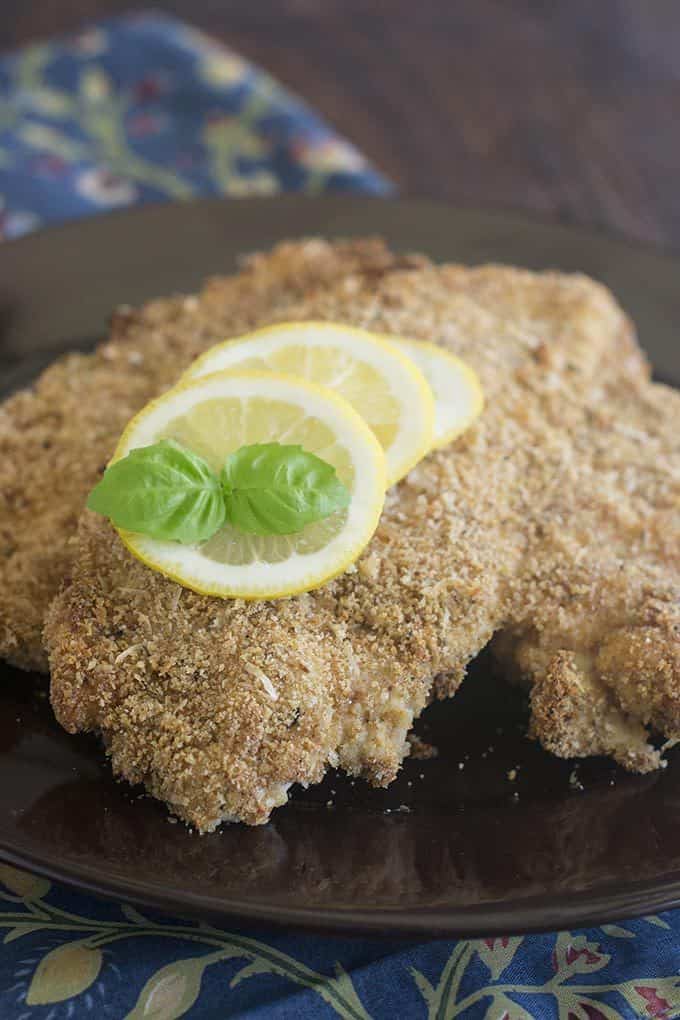 Close up of a flattened, breaded chicken breast that has been baked til golden brown and crispy. There are three lemon slices on top as well as a sprig from fresh basil. The chicken cutlet is sitting on a black, round plate on a blue floral cloth napkin.