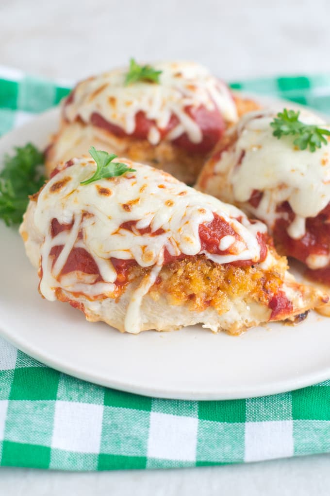 Three breaded chicken breasts on a round white plate are topped with red sauce, melted white cheese and garnished with parsley leaves. The plate is sitting on a green and white checked cloth napkin.