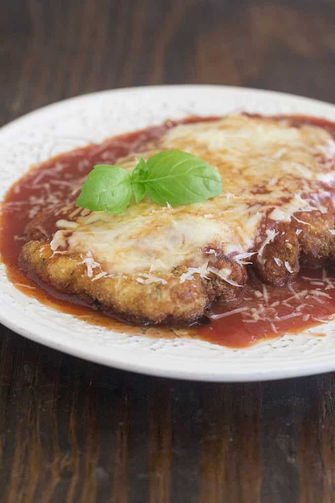 Chicken breast pounded flat, breaded and fried until golden and crispy. On a  round white plate smothered in red sauce and topped with melted white cheese. There is a sprig of fresh basil on top.
