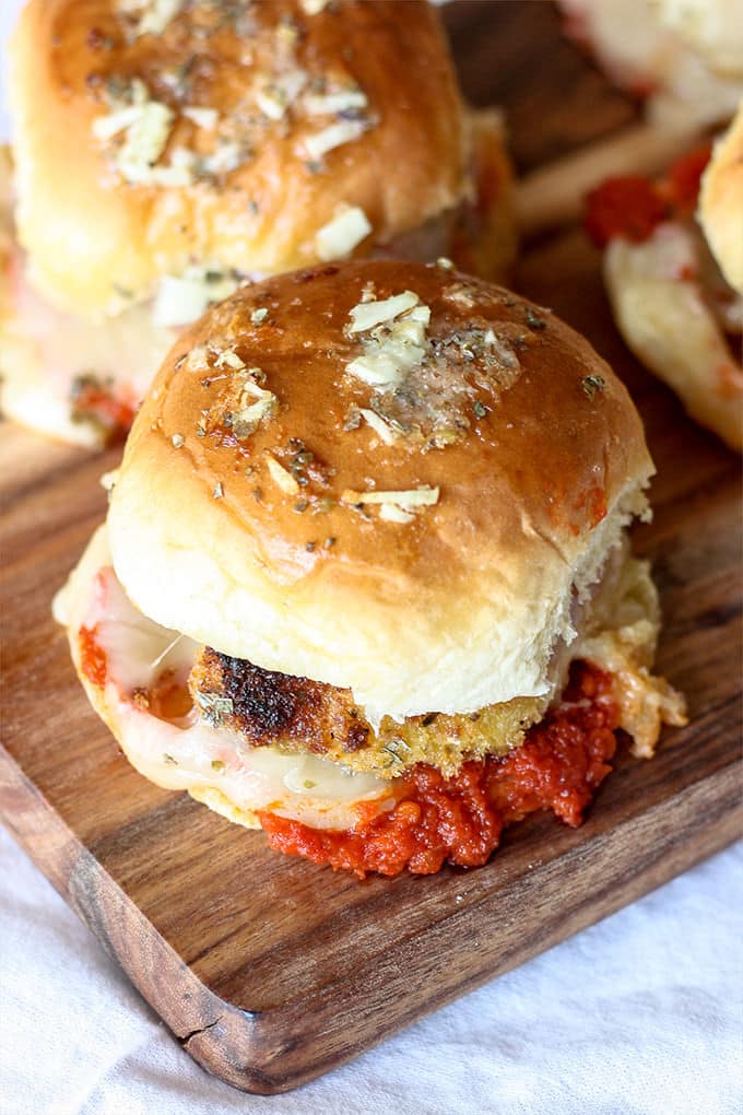 wooden cutting board with chicken parmesan sliders that have been brushed with melted butter with herbs and garlic. The slider bunds are filled with chicken parmesan, melted cheese and tomato sauce.