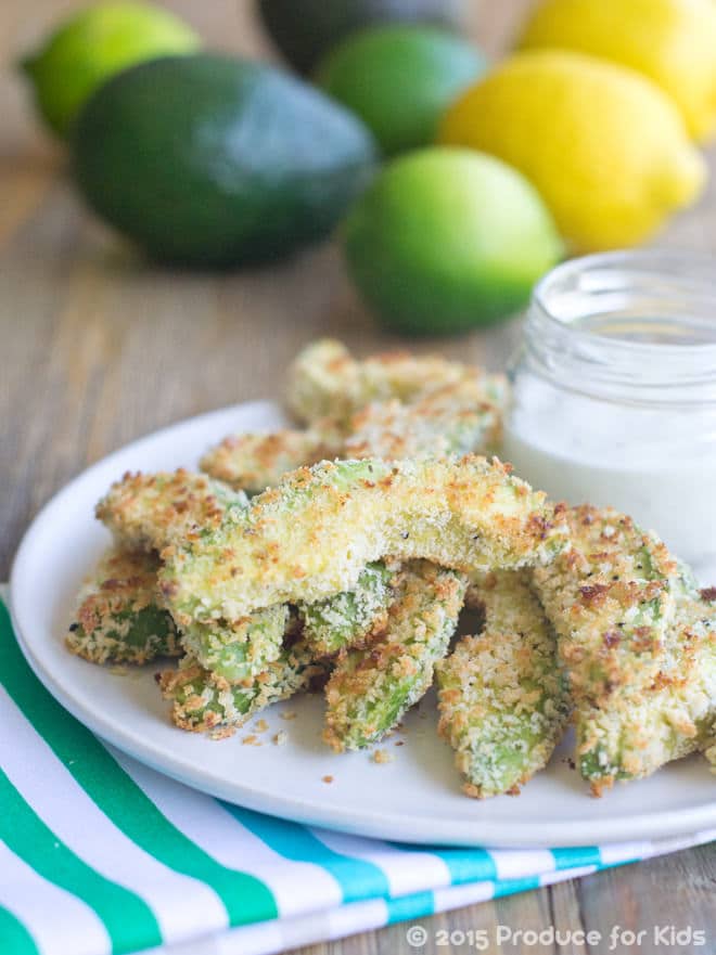 Breaded Avocado Fries on a white plate with a side of ranch dressing in a jar. Limes and Lemons in the background.