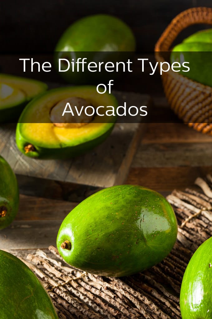 One half of an avocado with a pit on a cutting board with one whole avocado in the background as well as basket of avocados off to the side.