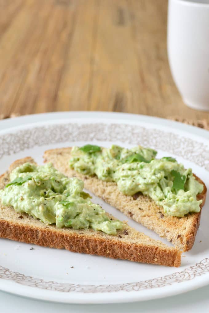 A piece of wheat toast on a plate, topped with smashed avocados, cilantro and pepper, sliced diagonally.