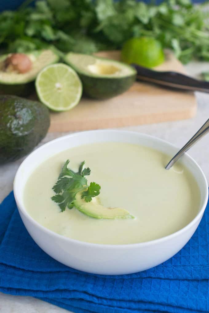 Bowl filled with avocado soup topped with a slice of avocado and sprig of cilantro. Halved avocado and half of lime and bunch of cilantro in background.