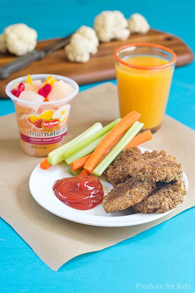 Small white plate with 4 cauliflower chicken nuggets, a dollop of ketchup and a pile of celery and carrot sticks.  The plate is sitting on a some brown butcher paper on a bright blue table. Also on the brown butcher paper is a glass of orange juice and cup of Del Monte mixed fruit. In the background is a wooden chopping board with some cauliflower florets and a knife.