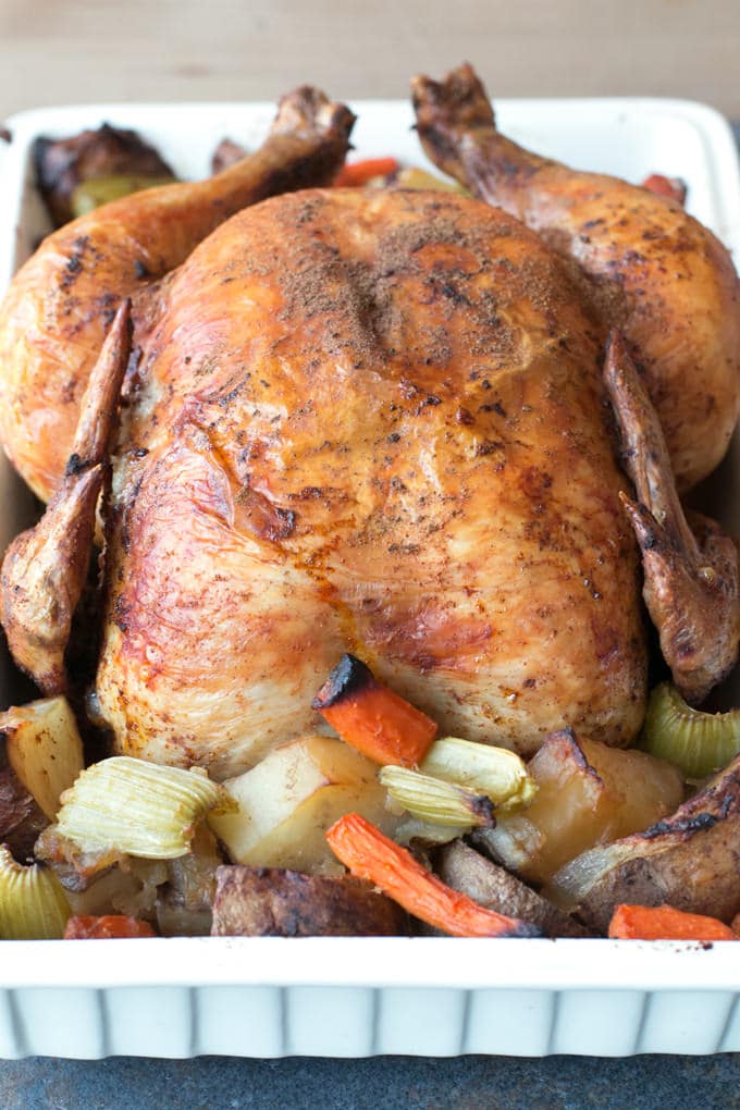 A whole chicken, cooked to a golden brown, in a white baking dish with roasted chopped potatoes, celery and carrots.