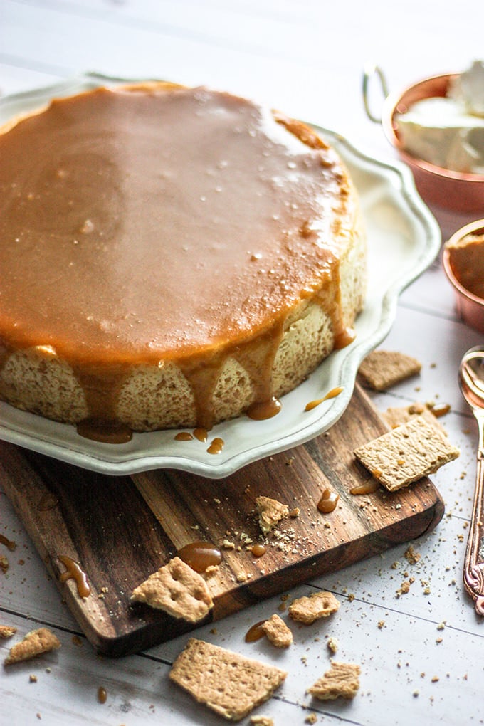 Whole cheese cake that has had caramel frosting poured over the top is sitting on a scalloped edge plate. The plate is sitting on a narrow wooden cutting board. The cutting board is on a white wood table that has been sprinkled with broken graham crackers and crumbs. In the background off to the right see a spoon, and a copper bowl of caramel and a copper bowl of cream cheese.