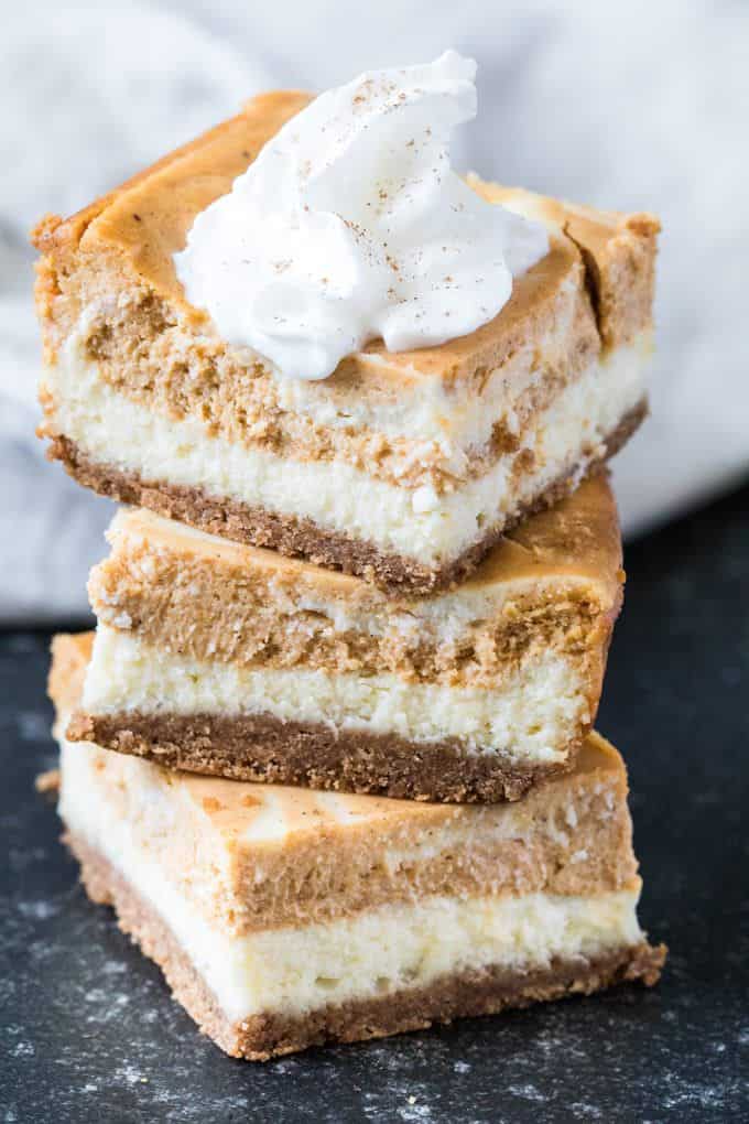 Three pumpkin cheesecake bars stacked on top of each other on a dark counter top. The top bar has a dollop of whipped cream on it.