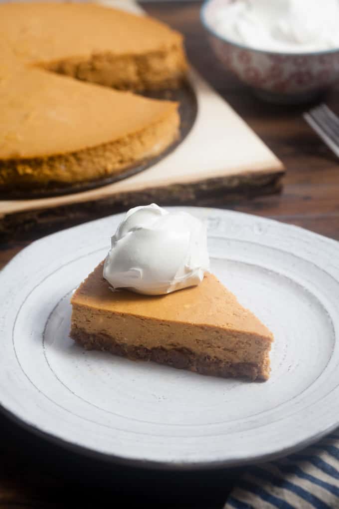 The Ultimate Pumpkin Cheesecake Recipe In the foreground is a large round white plate with a slice of pumpkin cheesecake topped with a dollop of whipped cream in the middle. In the background is a the whole cheesecake, with a piece missing, sitting on a wooden cutting board. Next to the cutting board is a red and white bowl filled with whipped cream.