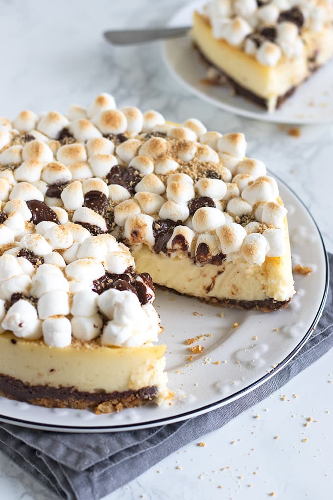 S'mores cheesecake topped with toasted mini marshmallows, drizzled with chocolate and sprinkled with graham cracker crumbs on a white plate with one piece missing. The plate is sitting on a grey cloth napkin on a white marble counter top that also has graham cracker crumbs sprinkled on it. In the background is a smaller white plate with a piece of the s'mores cheesecake as well as a fork.