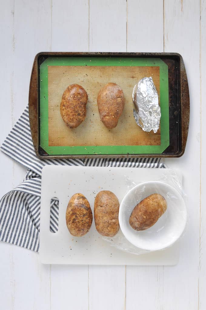 Metal cookie sheet with silicone baking mat has 3 whole baked potatoes (one wrapped in aluminum foil) sprinkled with salt and pepper and white plastic cutting board with 3 whole baked potatoes (one in a white bowl covered with plastic wrap)  sprinkled with salt and pepper are sitting on a blue and white stripped dish cloth on a white table.