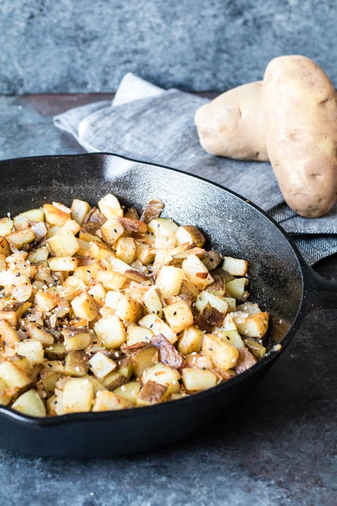 black cast iron skillet with cubed potatoes cooked in Homefry-style; behind the skillet is a black and white napkin with two whole russet potatoes on it. 