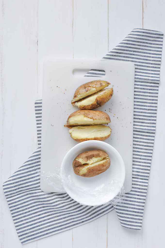 Three baked potatoes cut in half on a white cutting board, one in a white bowl with plastic wrap.