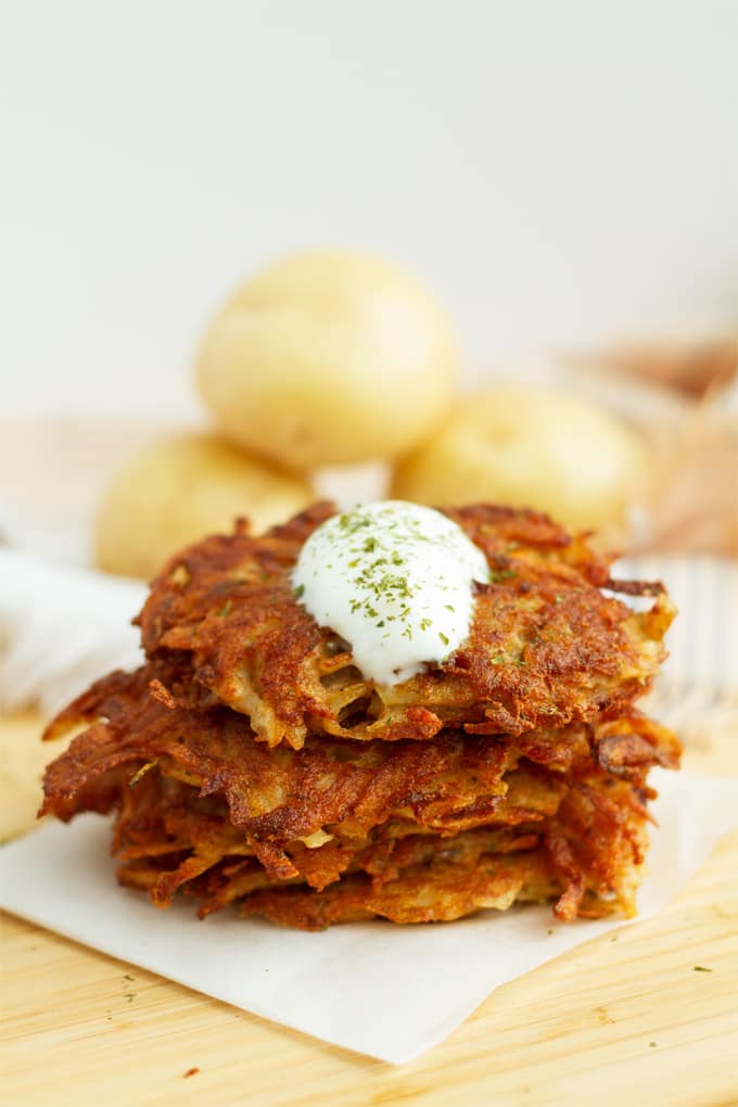 4 potato latkes stacked on top of each other on a piece of parchment paper on a wooden surface. The stack is topped with a dollop of sour cream. In the background you can see yellow potatoes.