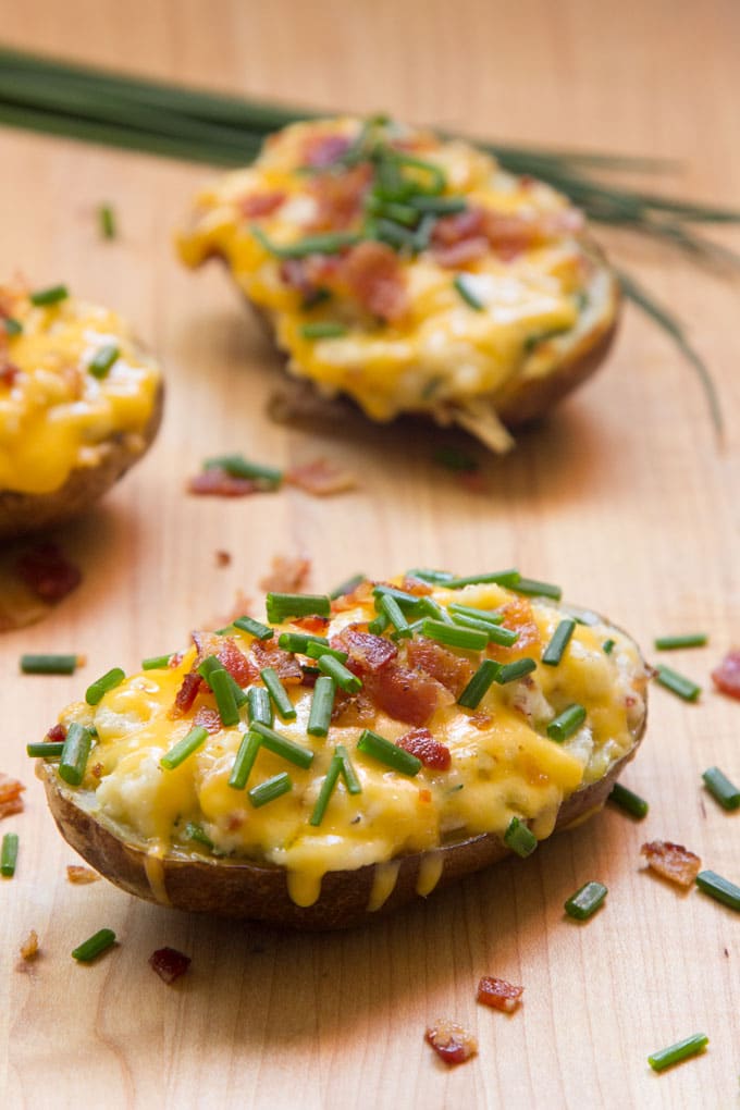 Two twice baked potato halves on a wooden cutting board topped with melted yellow cheddar cheese, chopped crispy bacon, and chives.