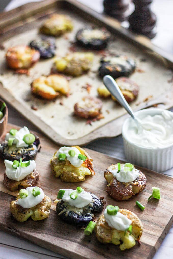 small, multicolored roasted and smashed potatoes on a long cutting board; potatoes are topped with a dollop of sour cream and chopped green onions; next to the cutting board is a small white ramekin; behind the cutting board is a baking sheet with roasted, smashed potatoes on it.