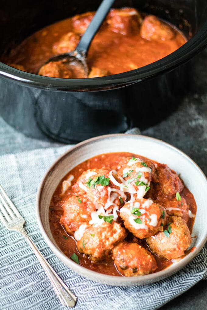 White bowl filled with meatballs in a red sauce stopped with shredded white cheese and garnished with diced parsley. In the background is a slow cooker filled with meatballs and red sauce.