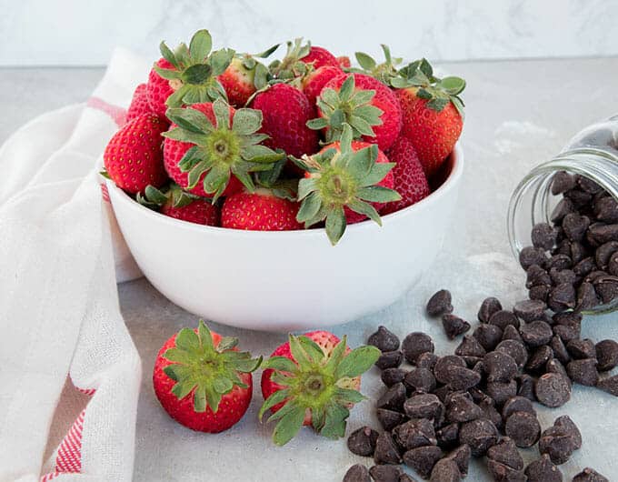Fresh strawberries in a white bowl with chocolate chips spilling out on the counter next to them.