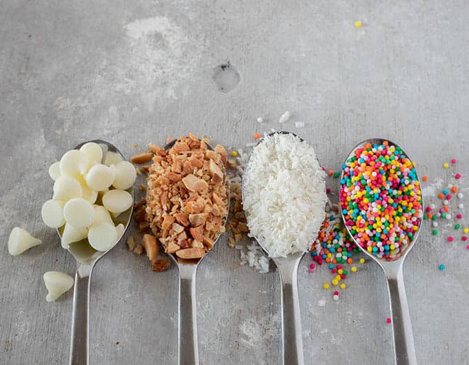 Spoons with various toppings - white chocolate, chopped nuts, coconut, and colorful sprinkles.