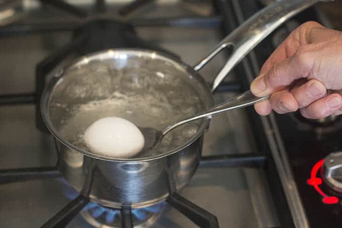 Egg being lowered into boiling water with a spoon.