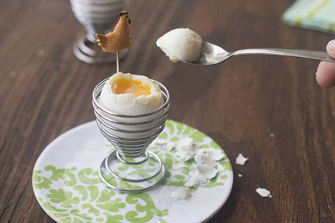 Spoon with cooked egg white top being lifted away from an egg in an egg cup, exposing the golden yolk.