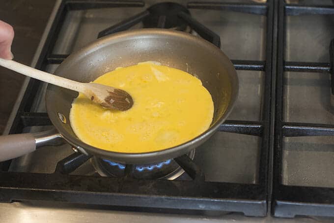 Whisked eggs in pan on stovetop being stirred with a wooden spoon.