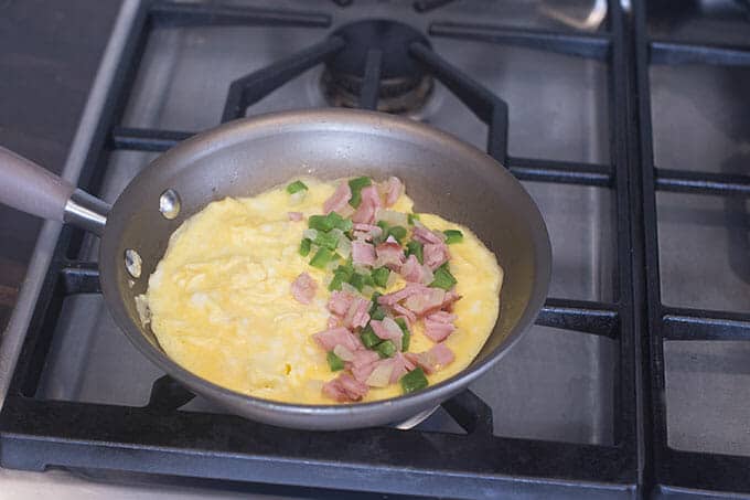 Eggs in a pan on the stove, with green pepper, onion, and ham topping one half.