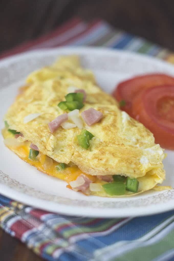 A Denver Omelet loaded with green bell pepper, onion, ham and cheddar cheese on a white plate with slices of tomato.