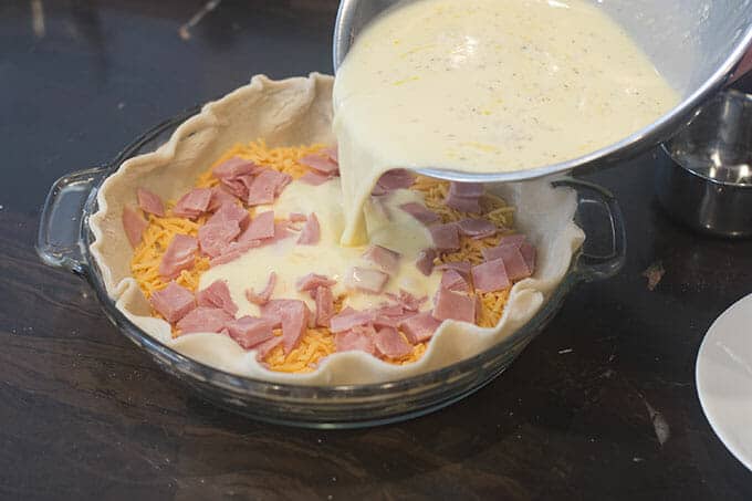 Egg mixture being poured over ham and cheese in pie crust.