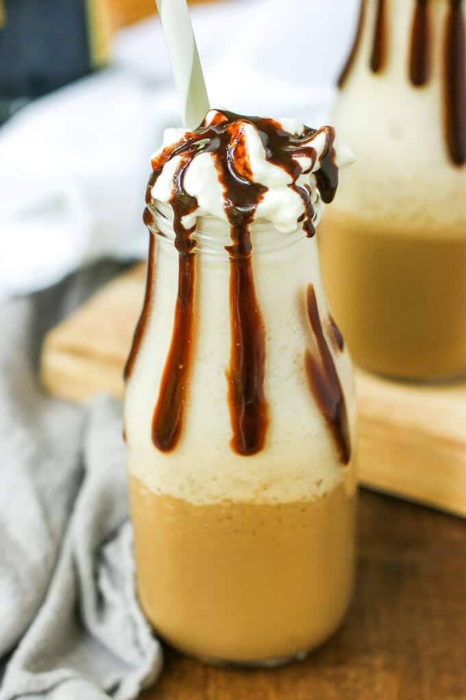 Small glass milk bottle filled with frozen fappuccino and whipped cream topped with chocolate sauce spilling over the sides with a straw stuck in the bottle. There is another similar bottle in the background.
