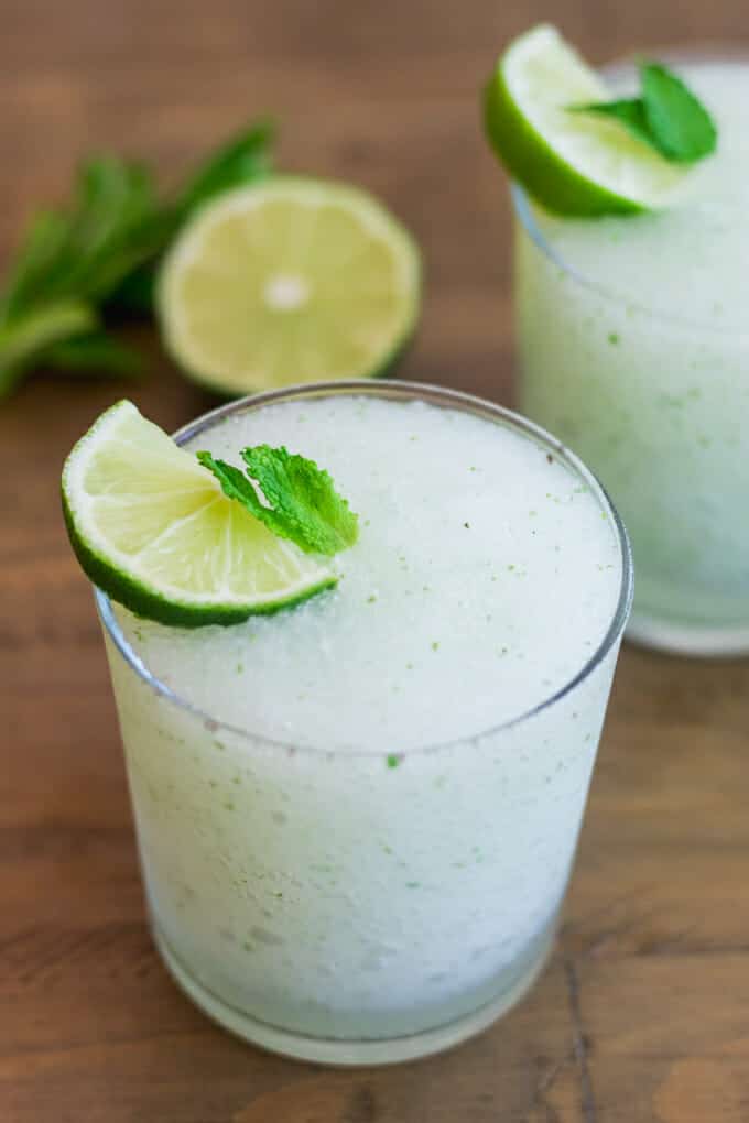 2 short glasses filled with frozen mojitos, topped with a slice of lime and small mint leaf. Glasses are sitting on a wooden surface with a half of a lime and some mint in the background.