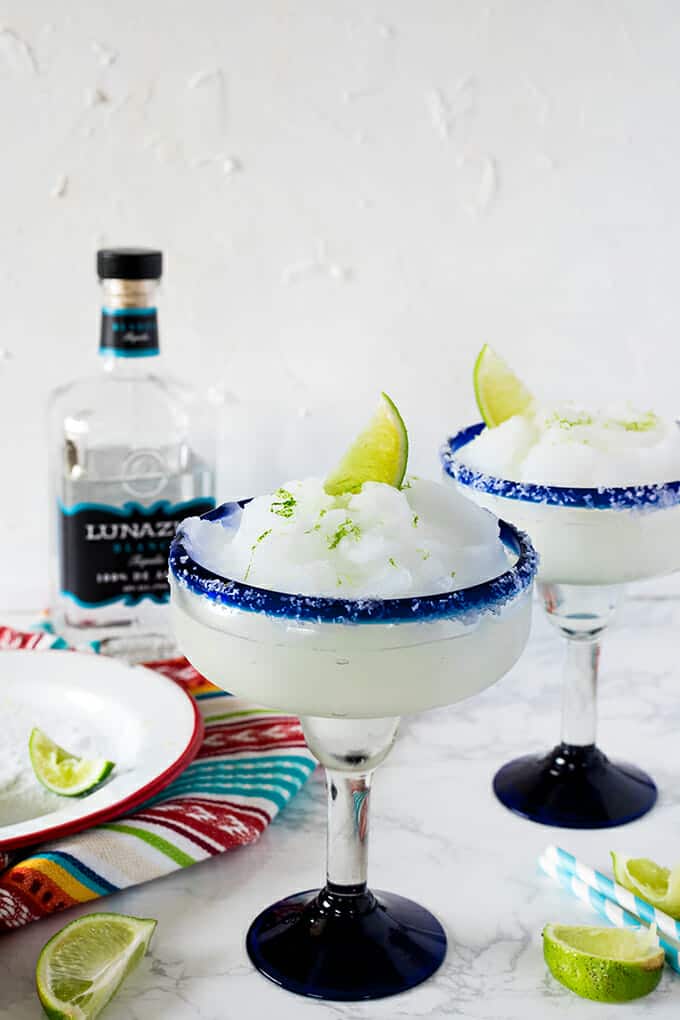 Two Margarita glasses filled with frozen Margarita and topped with a lime wedge and lime zest. The background has a bottle of tequila and a plate with salt and a lime wedge on a colorful dishcloth.