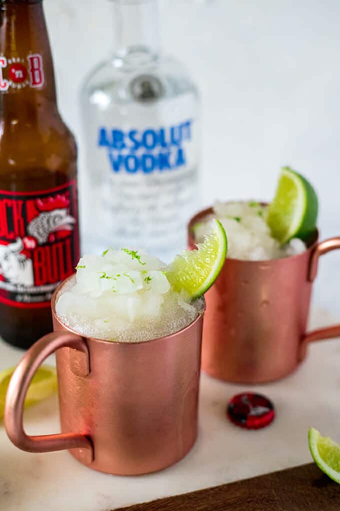 Two copper mugs filled with frozen Moscow Mule. Each mug is garnished with a wedge of lime and some lime zest. In the background is a bottle of Absolut vodka and bottle of ginger beer.