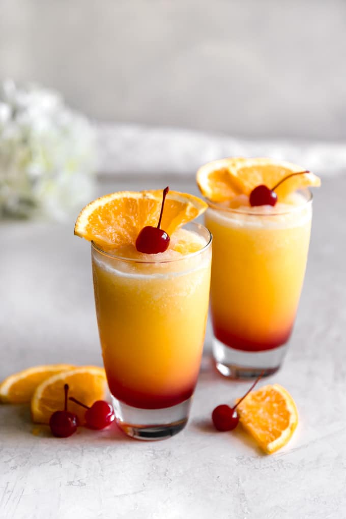 Two tall glasses filled with frozen Tequila Sunrise, which is red at the bottom and gradually transitions to yellow at the top. The glasses are garnished with a think orange lice and maraschino cherry and there are orange slices and cherries scattered around the glasses.