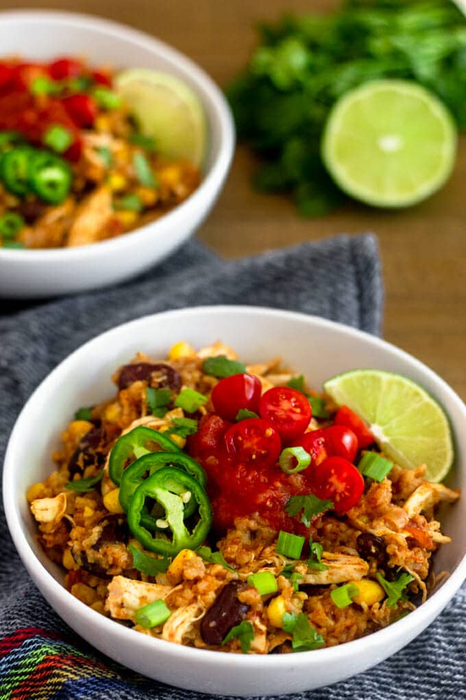 White bowl sitting on a black and white fabric placemat filled with rice cooked with bean and chicken topped with jalapeño slices, salsa, chopped tomatoes, a lime wedge and sprinkled with chopped green onions. In the background is an identical bowl, a half of a lime and a bunch of cilantro. 