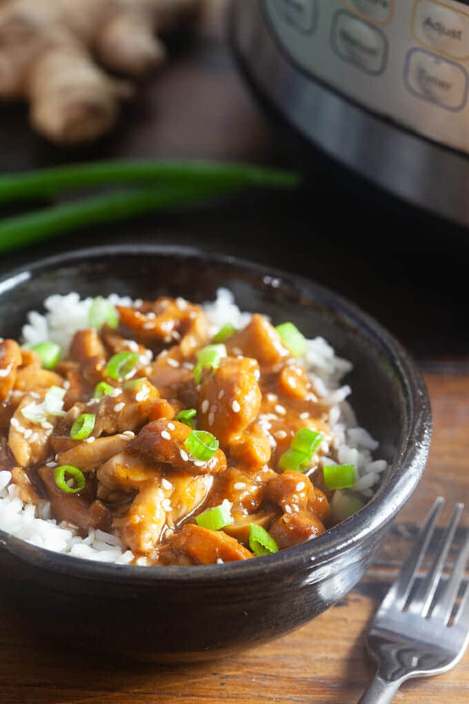 Black bowl filled with white rice topped with pieces of diced chicken in a brown sauce sprinkled with sesame seeds and diced green onions. There is a fork next to the bowl, and an instant pot, a bunch of green onions and a knob of ginger is in the background.