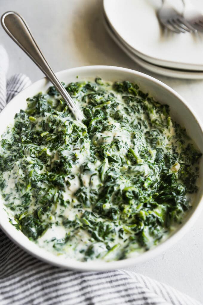 Creamed spinach in a white bowl with a silver spoon sticking out of it; in the back ground is a stack of plates with some silver forks on top.