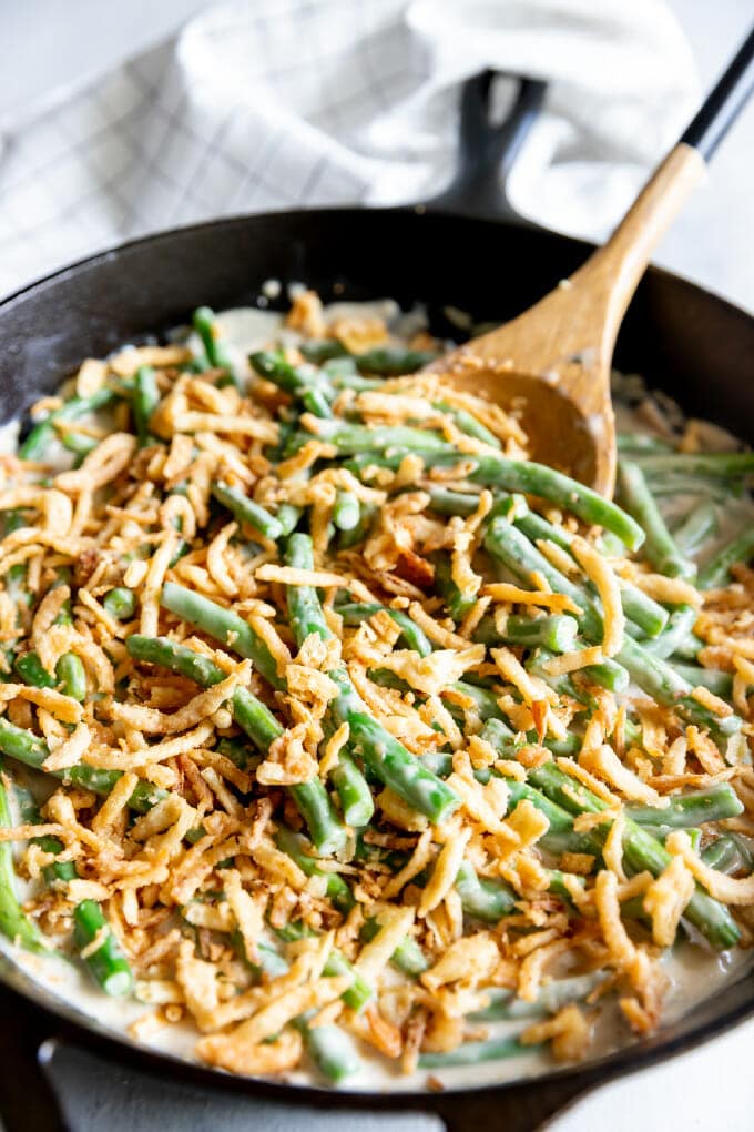 Green Bean Casserole topped with fried onions in a black cast iron skillet; there is a wooden spoon sticking out of the skillet.